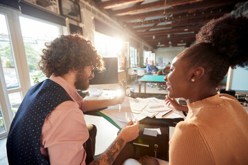 two young colleagues, male and female, different race, working together in the office, talking, smiling.