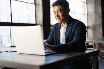 Wall Mural - Asian businessman typing on laptop during work in cafe. Concept of remote and freelance work. Smiling adult successful man wearing suit and glasses sitting at wooden desk. Sunny day