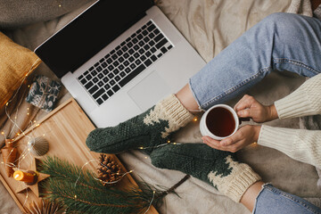 Wall Mural - Cozy winter holidays. Woman hands with cup of tea on bed with laptop, christmas lights, pine, candle