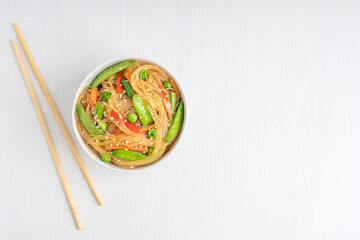 Wall Mural - Top view of Asian Japchae dish made of stir-fried glass noodles, green peas and beans with sesame seeds with savory flavor served in bowl with chopsticks on white wooden table. Image with copy space