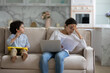 © fizkes - Tired young Indian freelance mom working from home, reading paper marketing report, documents on couch, holding laptop, watching little kid. Naughty kid distracting mum from distance work