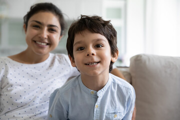 Happy Indian mom and preschool son looking at camera, speaking, laughing, sitting on couch at home. Little boy head shot with blurred mother in background. Video call screen view. Head shot portrait