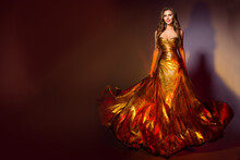 Fashion Model In Long Golden Dress. Smiling Beauty Woman In Evening Shining Fluttering Gold Gown Walking Over Dark Brown Background