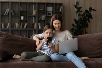 Cute girl child with smiling mother using gadgets at home together, sitting on couch, adorable little daughter with mom using modern smartphone and laptop, family having fun online, social media