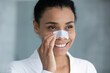 © fizkes - Head shot smiling attractive young african american biracial woman in bathrobe using nose cleansing strip, removing blackheads, enjoying skincare spa anti acne procedures, looking at camera.