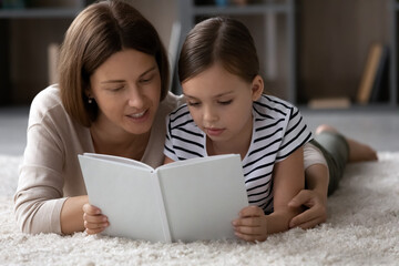 Wall Mural - Close up smiling mother with little girl reading book together, lying on warm floor at home, hugging, loving mom and adorable daughter kid learning, enjoying leisure time, educational activity