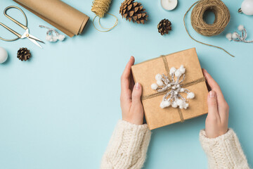 Wall Mural - First person top view photo of woman's hands in knitted pullover holding giftbox decorated with snow twig and twine christmas tree balls handicraft tools isolated pastel blue background with copyspace