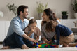 © fizkes - Happy parents and two little kids playing with toys on warm floor carpet, having fun together, laughing mother and father in glasses with children building colorful plastic constructor blocks