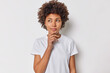 © wayhome.studio  - Attractive curly haired woman keeps hand on chin considers something and looks away imagines smth in mind makes has idea wears casual t shirt isolated over white background. Thoughts concept