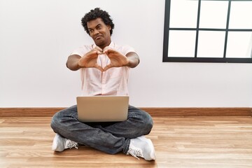 Canvas Print - African man with curly hair using laptop sitting on the floor smiling in love doing heart symbol shape with hands. romantic concept.