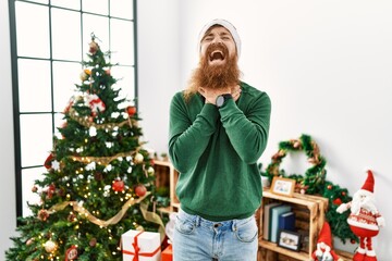 Poster - Redhead man with long beard wearing christmas hat by christmas tree shouting suffocate because painful strangle. health problem. asphyxiate and suicide concept.