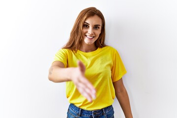 Poster - Young brunette woman standing over isolated background smiling friendly offering handshake as greeting and welcoming. successful business.