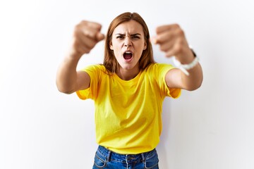 Wall Mural - Young brunette woman standing over isolated background angry and mad raising fists frustrated and furious while shouting with anger. rage and aggressive concept.