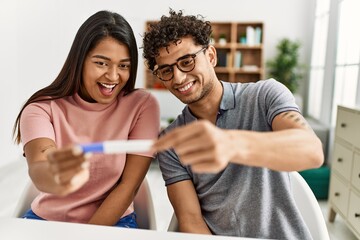 Young latin couple smiling happy holding pregnancy test sitting on the table at home.