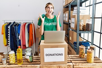 Wall Mural - Young blonde woman wearing volunteer t shirt at donations stand crazy and mad shouting and yelling with aggressive expression and arms raised. frustration concept.