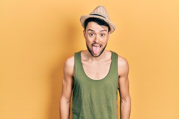 Poster - Young hispanic man wearing summer hat sticking tongue out happy with funny expression. emotion concept.