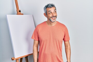 Canvas Print - Handsome middle age man with grey hair standing by painter easel stand smiling looking to the side and staring away thinking.