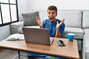 Sticker - Young handsome hispanic man using laptop sitting on the floor crazy and mad shouting and yelling with aggressive expression and arms raised. frustration concept.