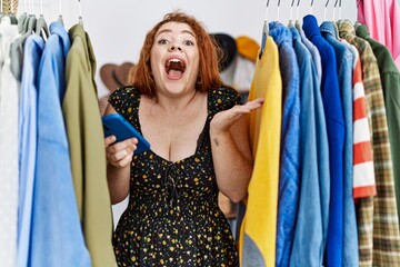 Wall Mural - Young redhead woman searching clothes on clothing rack using smartphone celebrating victory with happy smile and winner expression with raised hands