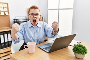 Canvas Print - Young redhead woman working at the office using computer laptop angry and mad raising fists frustrated and furious while shouting with anger. rage and aggressive concept.