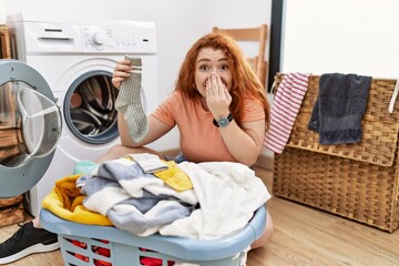 Wall Mural - Young redhead woman putting dirty laundry into washing machine laughing and embarrassed giggle covering mouth with hands, gossip and scandal concept