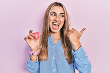 Sticker - Beautiful hispanic woman holding menstrual cup pointing thumb up to the side smiling happy with open mouth