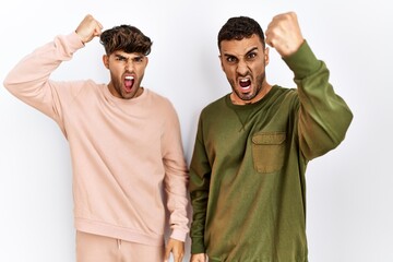 Poster - Young gay couple standing over isolated white background angry and mad raising fist frustrated and furious while shouting with anger. rage and aggressive concept.