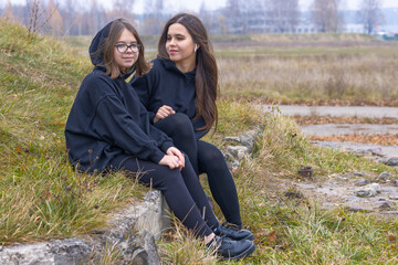 Two beautiful girls with long hair in black clothes sit together on the grass on the outskirts of the city.