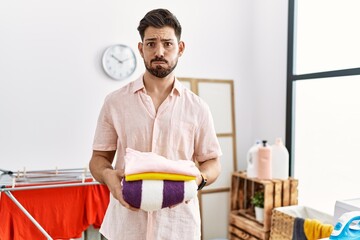 Poster - Young man with beard holding folded laundry after ironing puffing cheeks with funny face. mouth inflated with air, catching air.