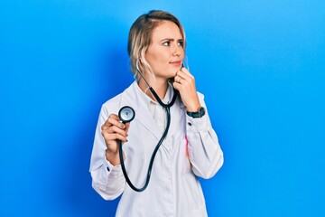 Wall Mural - Beautiful young blonde doctor woman holding stethoscope with hand on chin thinking about question, pensive expression. smiling with thoughtful face. doubt concept.