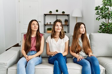 Poster - Group of three hispanic girls sitting on the sofa at home smiling looking to the side and staring away thinking.