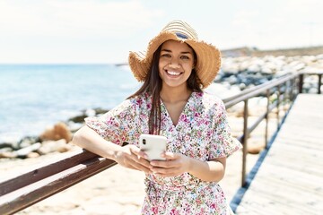 Sticker - Young latin girl wearing summer hat using smartphone at the beach.