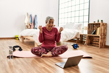 Poster - Middle age woman with grey hair training at home looking at exercise video on laptop celebrating mad and crazy for success with arms raised and closed eyes screaming excited. winner concept