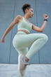 Vertical shot of sporty female runner warms up outdoors stretches legs keeps knees raised dressed in activewear and sportshoes poses against concrete grey wall exercises outdoors gets ready for run