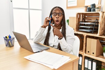 Poster - Black woman with braids working at the office speaking on the phone surprised pointing with finger to the side, open mouth amazed expression.