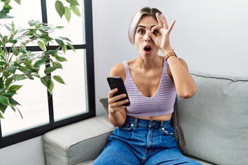 Sticker - Young beautiful woman using smartphone typing message sitting on the sofa doing ok gesture shocked with surprised face, eye looking through fingers. unbelieving expression.