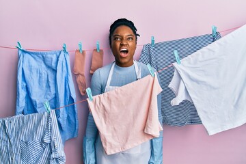 Wall Mural - African american woman with braided hair washing clothes at clothesline angry and mad screaming frustrated and furious, shouting with anger. rage and aggressive concept.