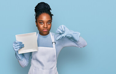 Poster - Young african american woman wearing apron washing dishes with angry face, negative sign showing dislike with thumbs down, rejection concept