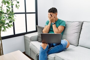 Wall Mural - Young handsome man with beard using computer laptop sitting on the sofa at home with sad expression covering face with hands while crying. depression concept.