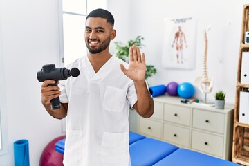 Sticker - Young indian physiotherapist holding therapy massage gun at wellness center waiving saying hello happy and smiling, friendly welcome gesture