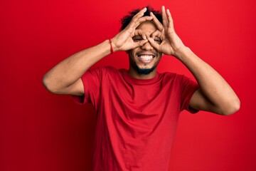 Poster - Young african american man with beard wearing casual red t shirt doing ok gesture like binoculars sticking tongue out, eyes looking through fingers. crazy expression.