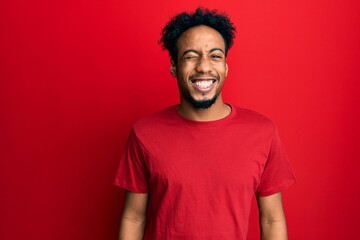 Wall Mural - Young african american man with beard wearing casual red t shirt winking looking at the camera with sexy expression, cheerful and happy face.