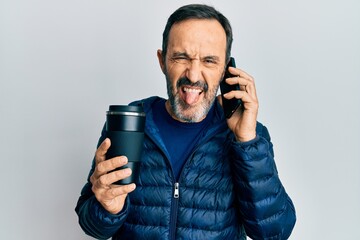 Poster - Middle age hispanic man talking on the smartphone and drinking a cup of coffee sticking tongue out happy with funny expression.