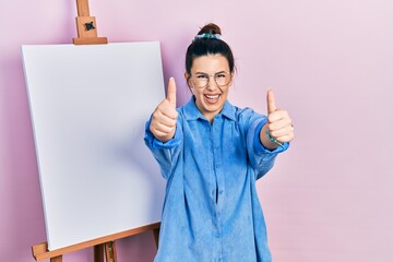 Poster - Young hispanic woman standing by painter easel stand approving doing positive gesture with hand, thumbs up smiling and happy for success. winner gesture.