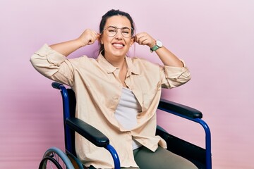 Sticker - Young hispanic woman sitting on wheelchair smiling pulling ears with fingers, funny gesture. audition problem