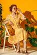 Fashionable redhead freckled woman wearing trendy summer midi dress, yellow square sunglasses, with classic leather bag, posing on yellow background. Full-length portrait