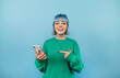 © bodnarphoto - Positive bright lady in a green sweatshirt and colored hair stands on a blue background with a smartphone in his hands and looks at the camera with a smile on his face.