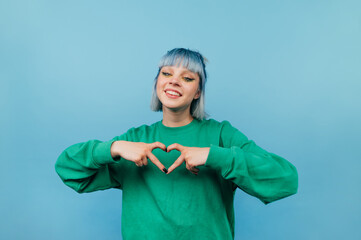 Wall Mural - Positive hipster girl with dyed hair shows a heart gesture to the camera those looking with a smile on her face.