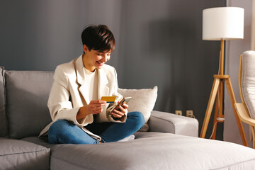 Young woman sits on a sofa shopping over a smartphone and uses a credit card