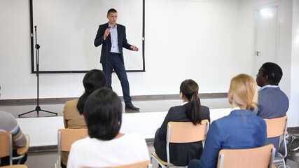 Wall Mural - Confident lecturer standing with microphone on stage in conference room, speaking to businesspeople at seminar